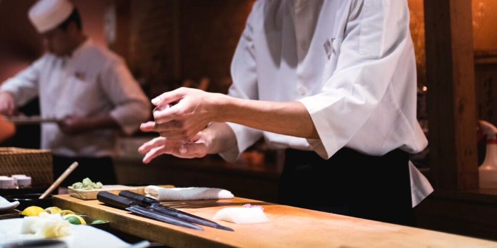 Indoor photo of an open bar sushi restaurant with two Japanese chefs making sushi, one in foreground and one in the background.